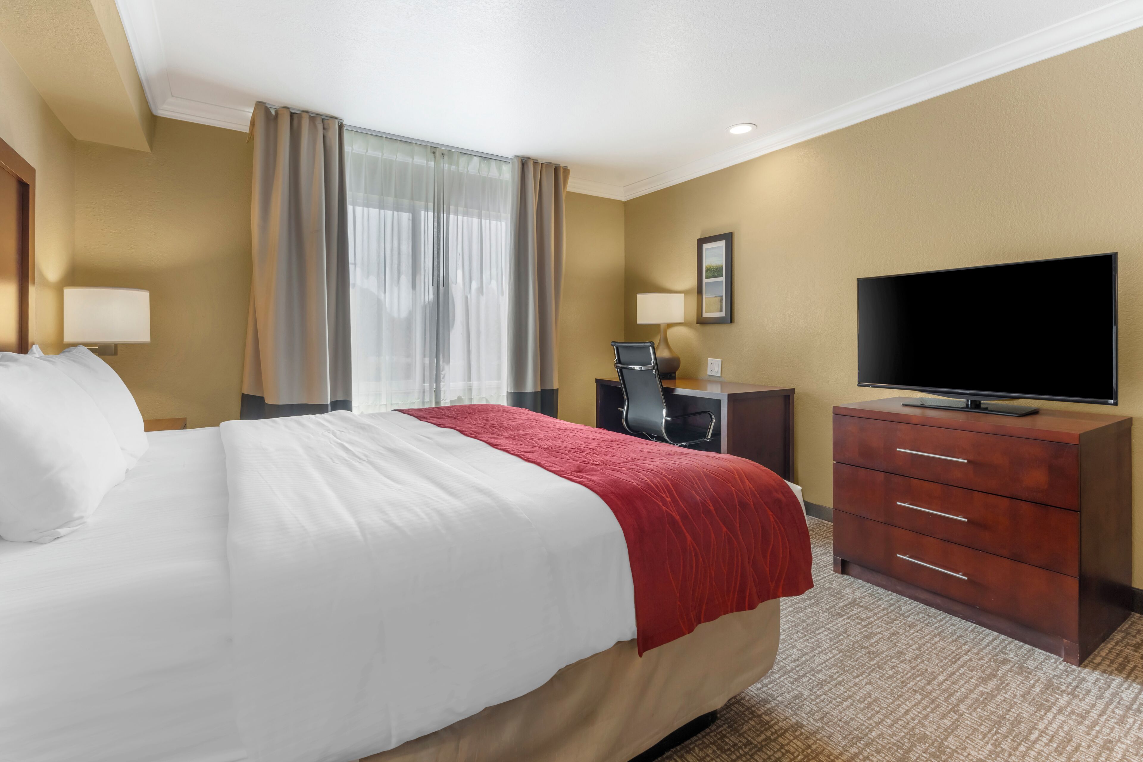 A hotel room with a large bed, a desk with a chair, a television, and a dresser.