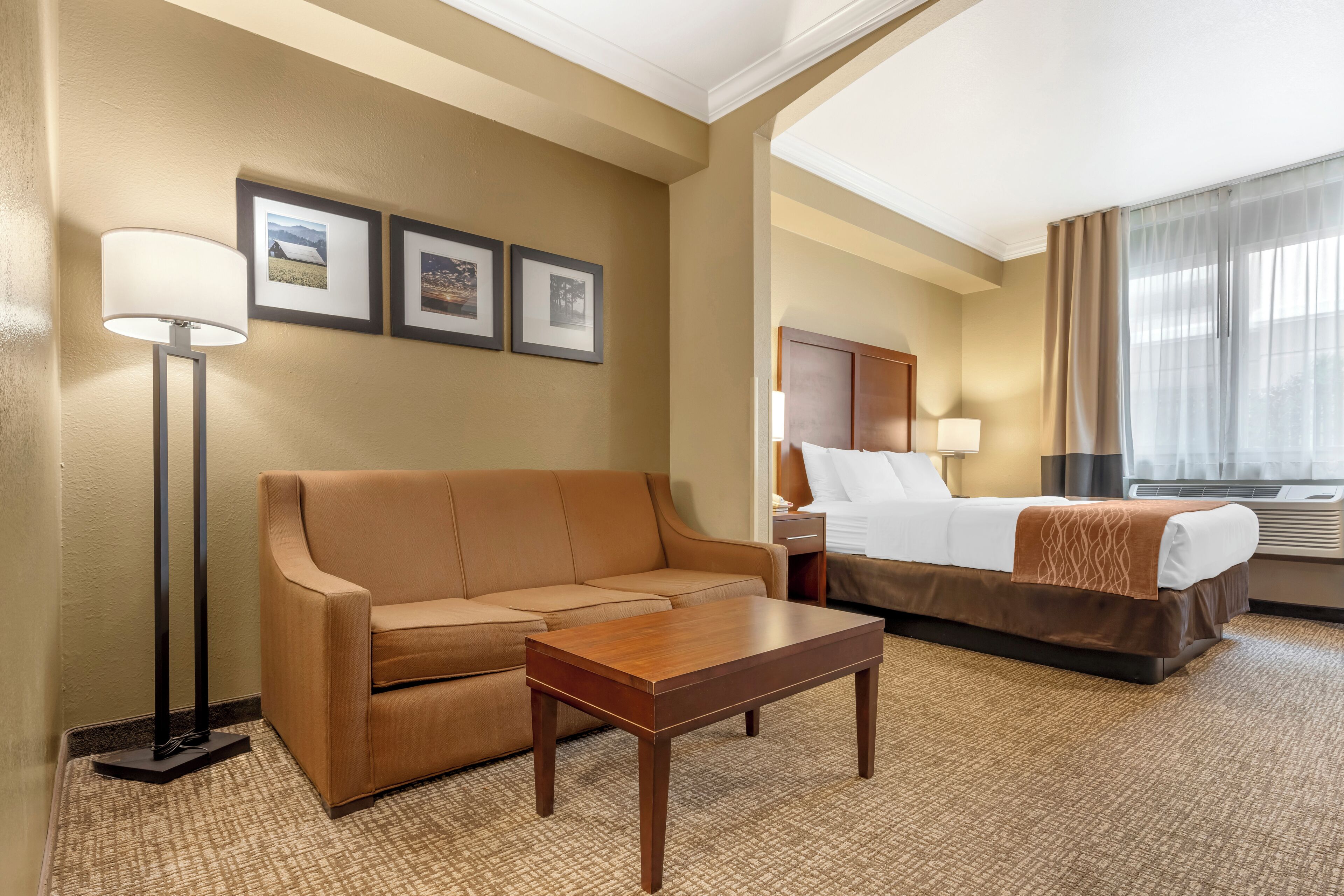 A hotel room with a sofa, a wooden table, a bed with a brown bedspread, a nightstand with a lamp, and framed pictures on the wall.