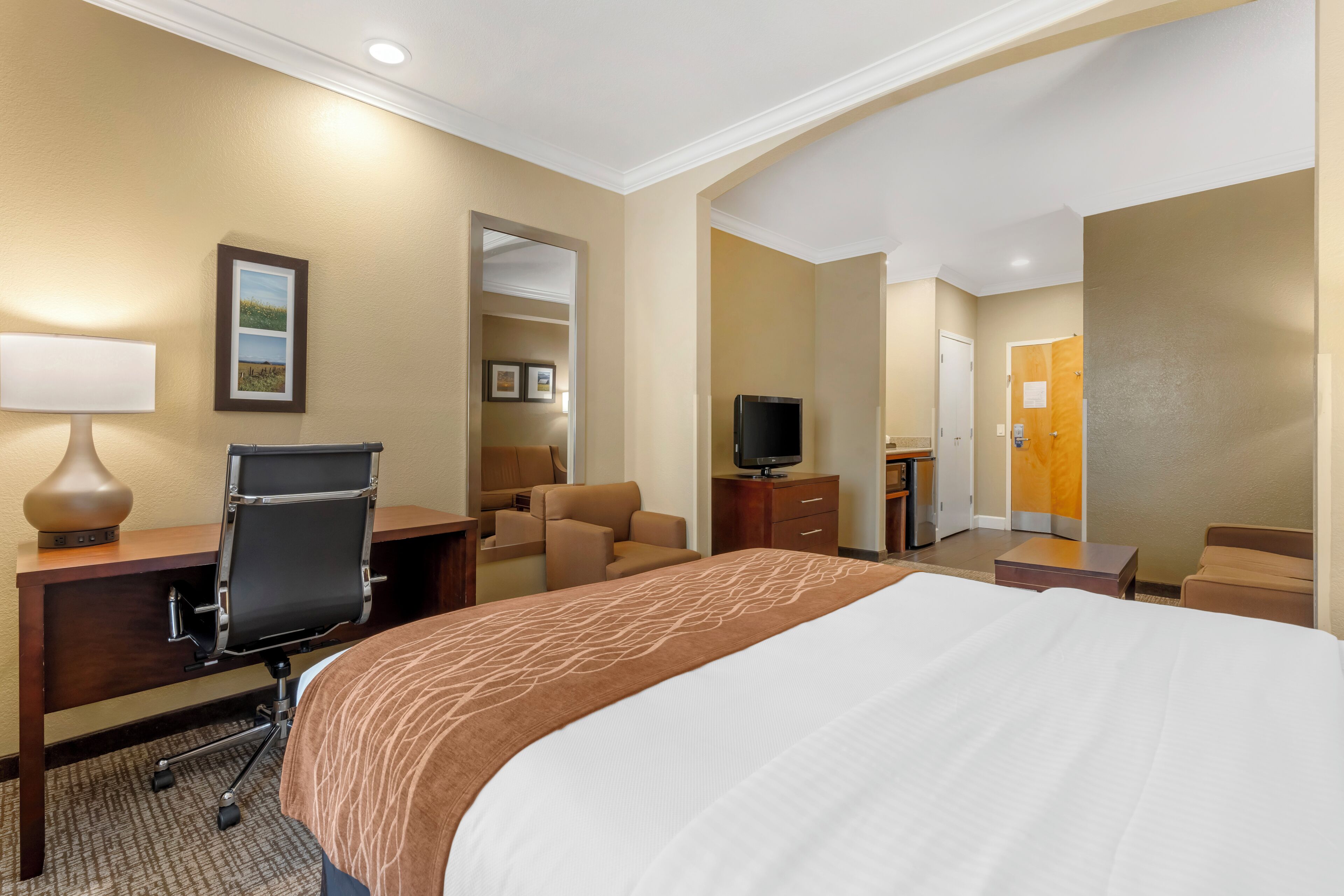 A hotel room with a large bed, a desk with a chair, a television, and a bathroom visible through an open door.
