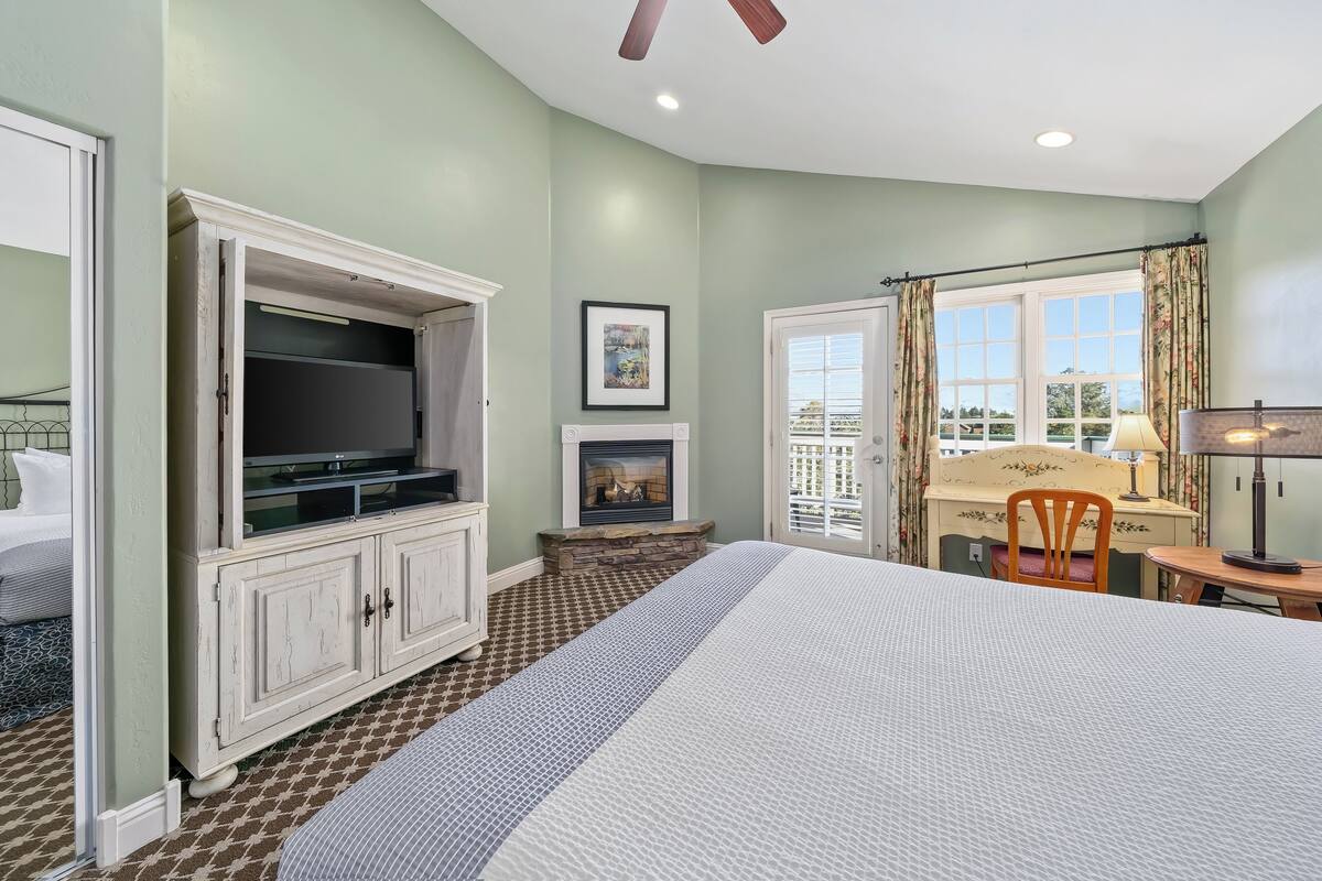 A bedroom with a bed, a television, a fireplace, a desk with a chair, and a window with curtains.