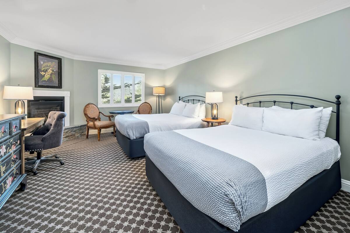 A hotel room with two beds, a desk with a chair, a fireplace, and a window with a view.
