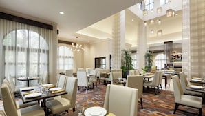A spacious hotel dining area with multiple tables and chairs, large windows, and a high ceiling with hanging lights. - Hilton Garden Inn Cupertino (Cupertino)