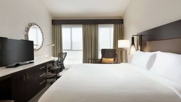 A hotel room with a large bed, a TV, a desk with drawers, a chair, a lamp, and a window with curtains.