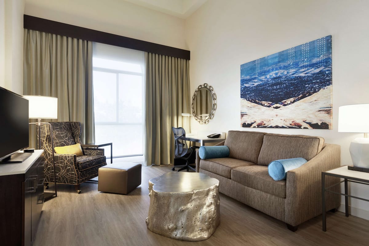 A modern hotel room with a large window, a sofa, a desk, and a television.