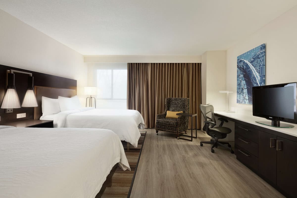 A hotel room with two beds, a desk with a television, a chair, and a large window with curtains.