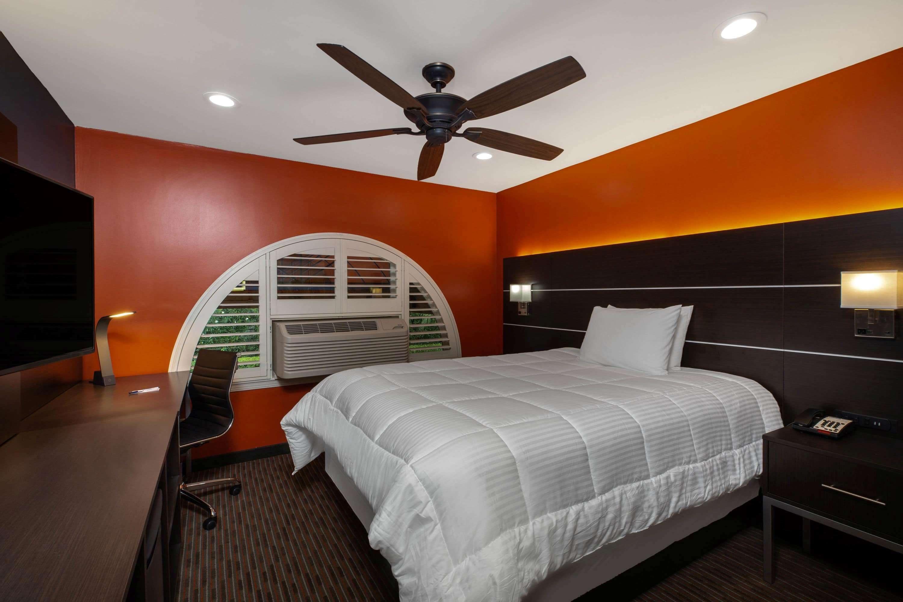 Quarto de hotel com cama, escrivaninha, cadeira, ventilador de teto, televisão na parede e janela com persianas.