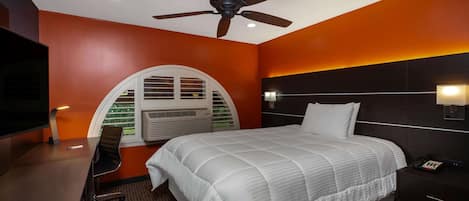 Une chambre d’hôtel équipée d’un lit, d’un bureau, d’une chaise, d’un ventilateur de plafond, d’un téléviseur fixé au mur et d’une fenêtre avec des stores.