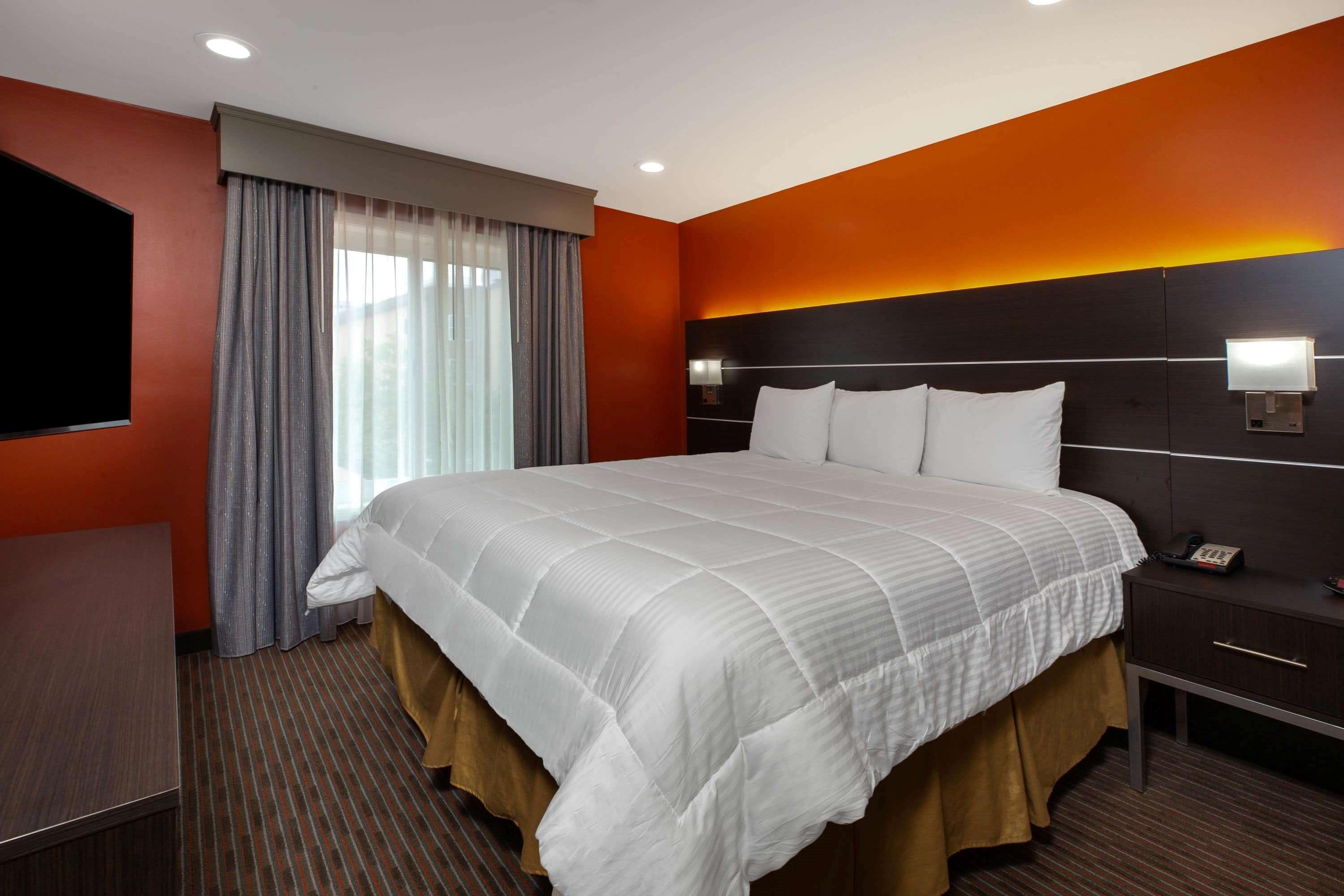 Habitación de hotel con una cama grande, una mesita de noche con lámpara, un televisor montado en la pared y una ventana con cortinas.