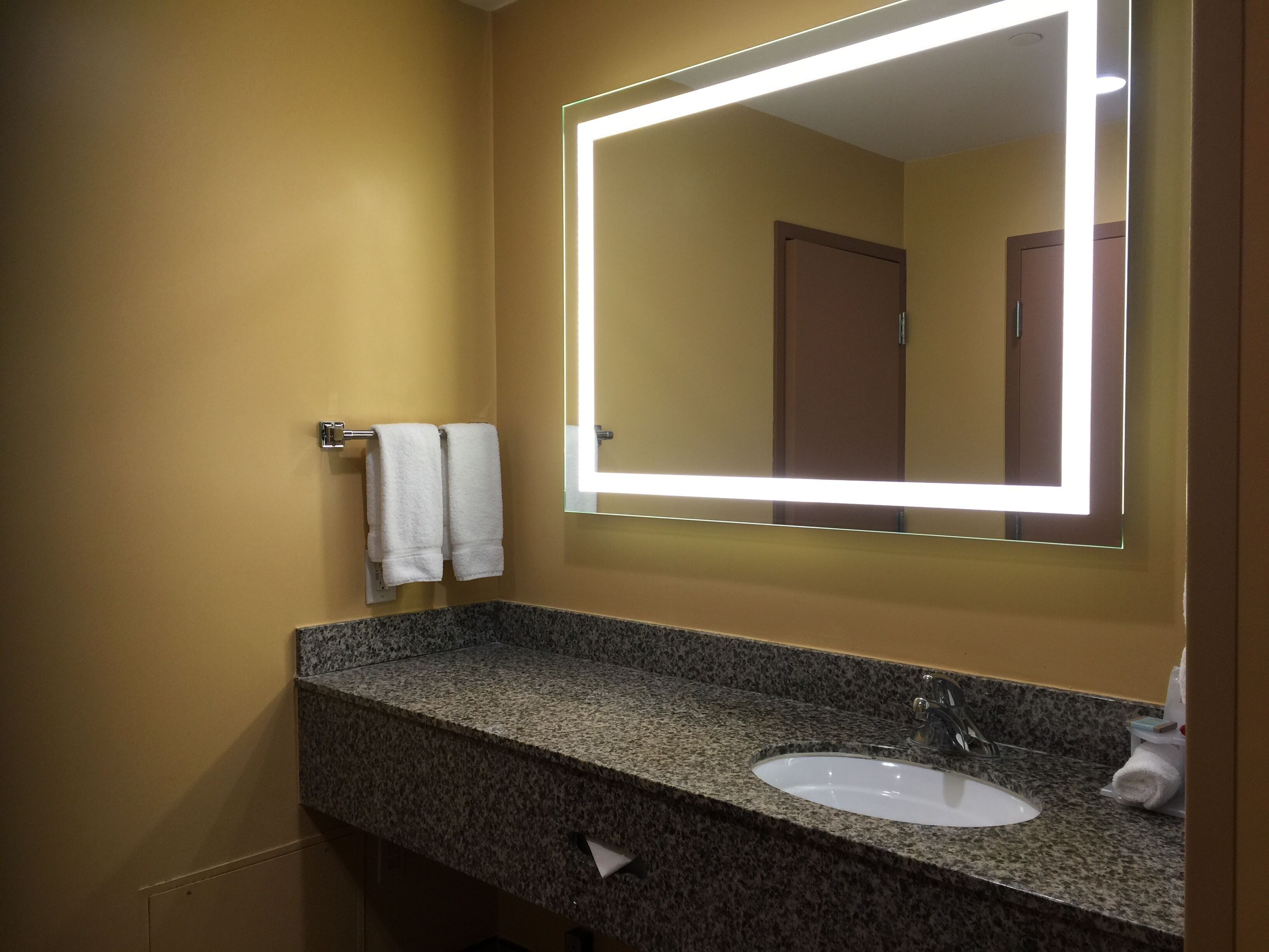 A bathroom with a granite countertop, a white sink, and a mirror with a lighted frame.
