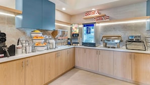 A modern kitchen with a breakfast bar, a coffee machine, a fruit bowl, and a sign that reads "RISE & SHINE".
