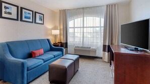 A hotel room with a blue sofa, a wooden desk, a flat-screen TV, a window with curtains, and framed pictures on the wall.