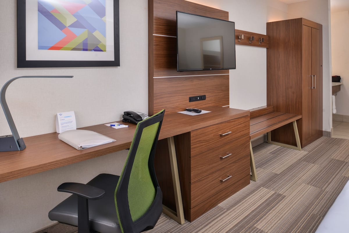 Ein Hotelzimmer mit Schreibtisch aus Holz, einem darüber montierten Fernseher, einem grünen Bürostuhl und einem Kleiderschrank.