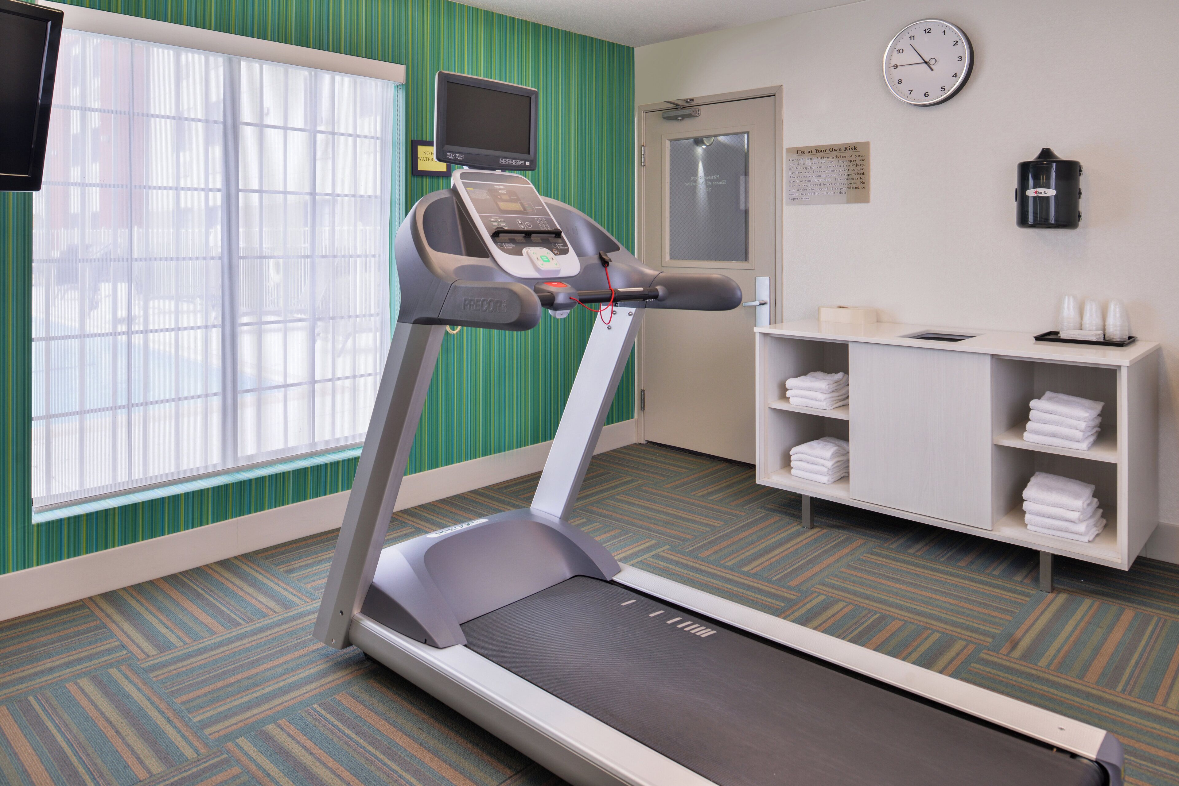 A hotel room with a treadmill, a clock, a TV, and a shelf with towels.