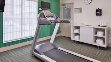 A hotel room with a treadmill, a clock, a TV, and a shelf with towels.