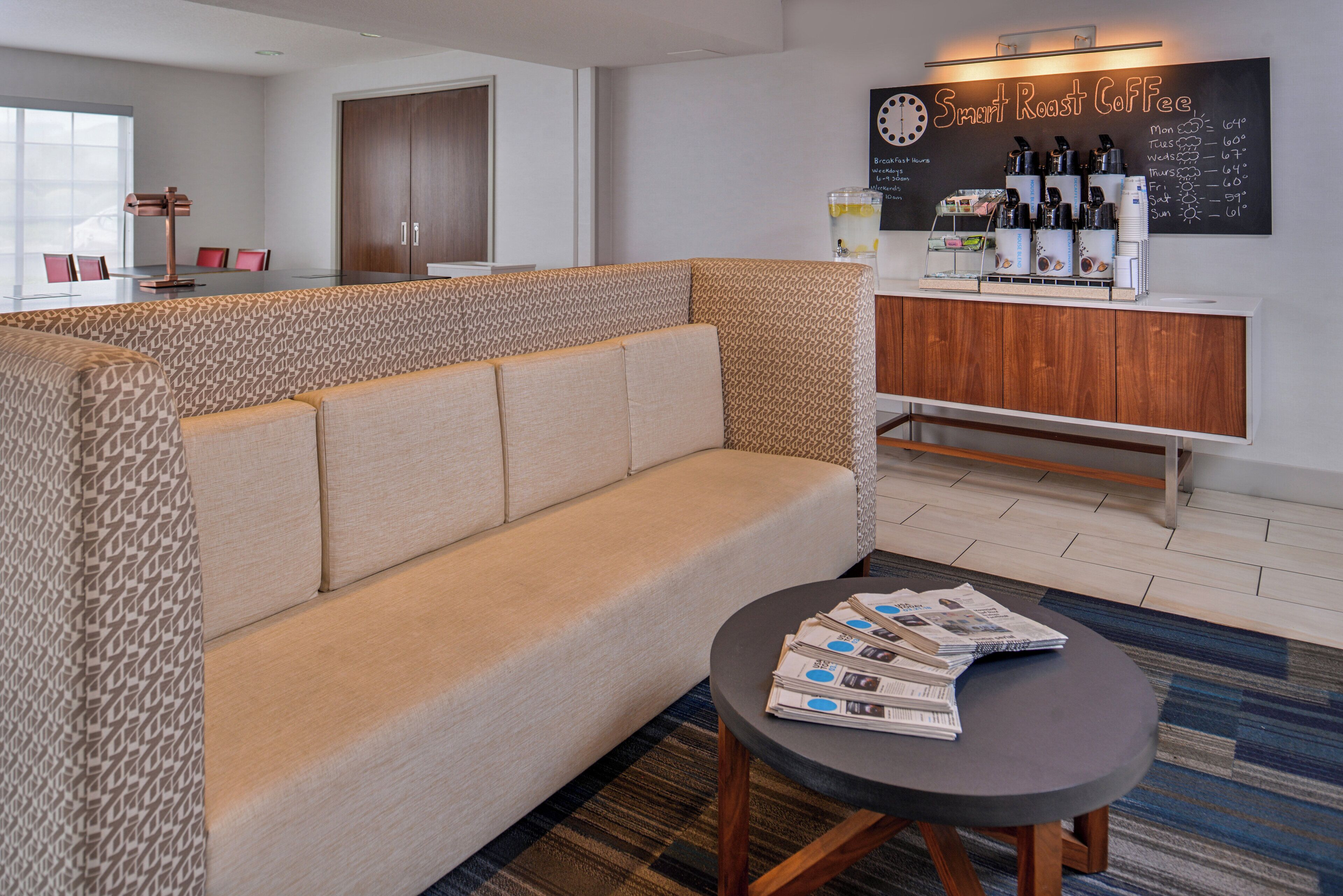 A modern lounge area with a sofa, a small table with newspapers, and a coffee station with a chalkboard displaying the weather.