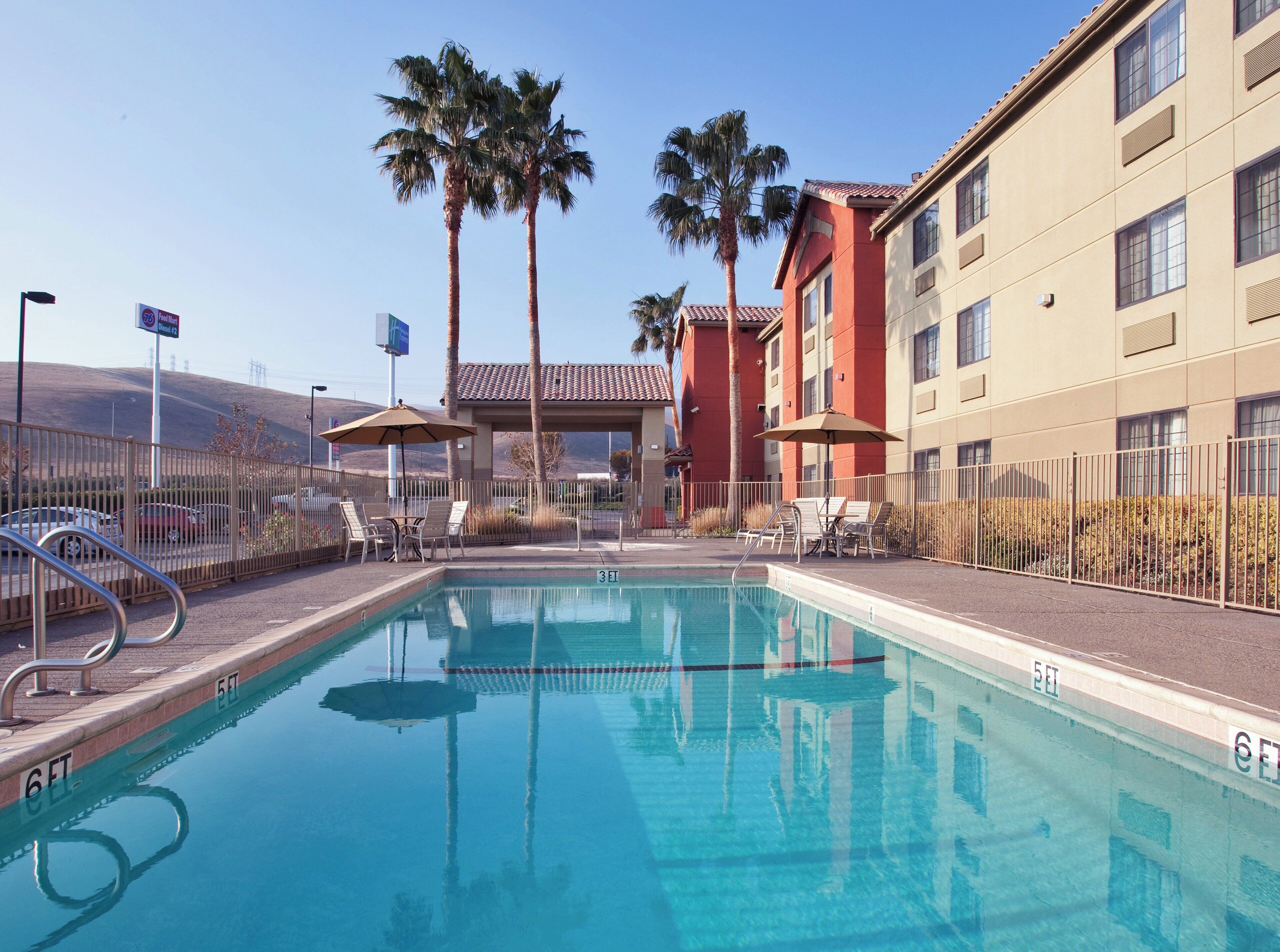 A swimming pool with lounge chairs and umbrellas, surrounded by a fence, with a building in the background.