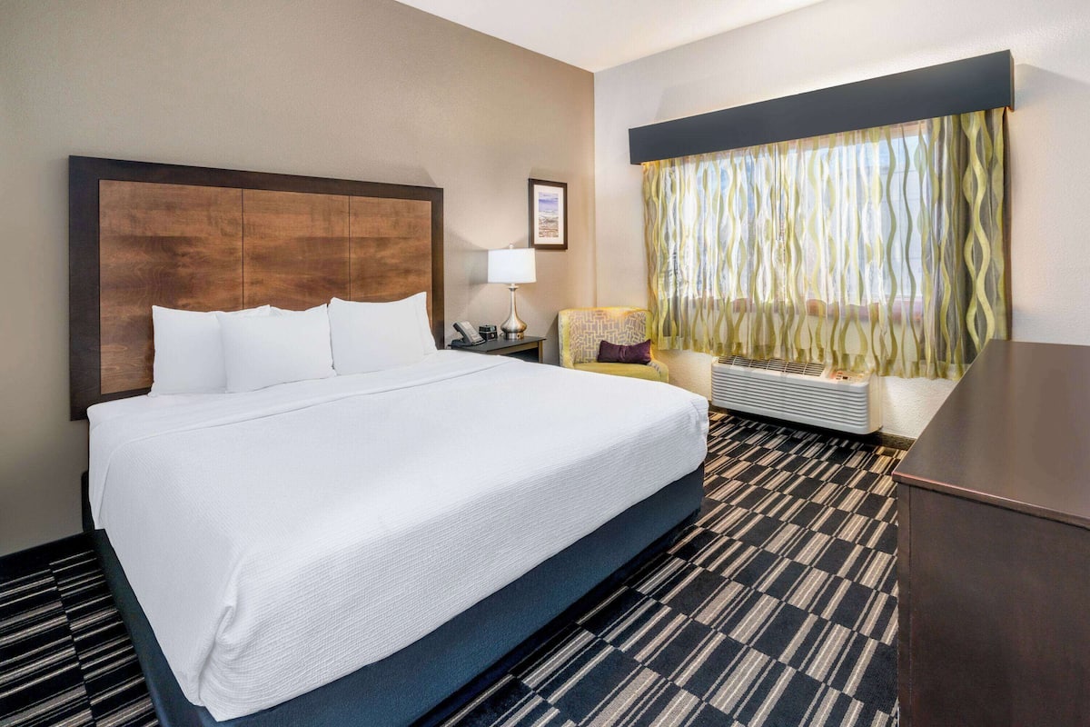 Een hotelkamer met een groot bed, een nachtkastje met een lamp, een stoel, een raam met gordijnen, en een ingebouwde heater.
