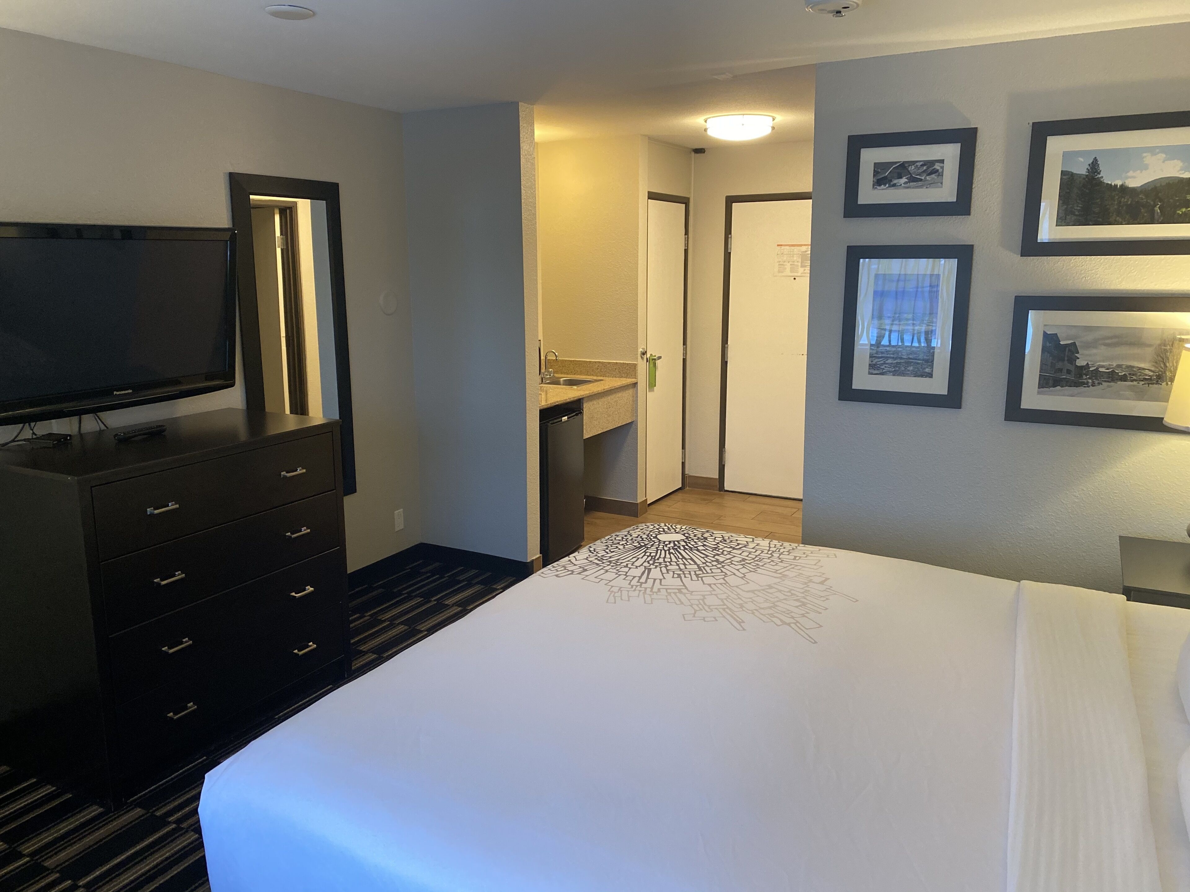 A hotel room with a bed, a TV, a dresser, a bathroom, and framed pictures on the wall.