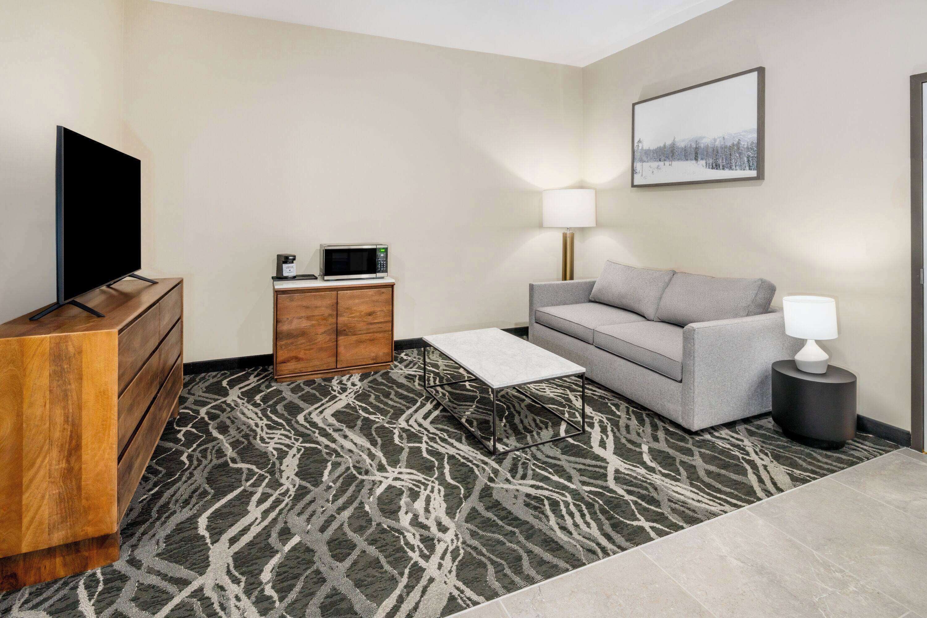 A hotel room with a flat-screen TV, a wooden dresser, a gray sofa, a small table with a lamp, a microwave, and a patterned rug.