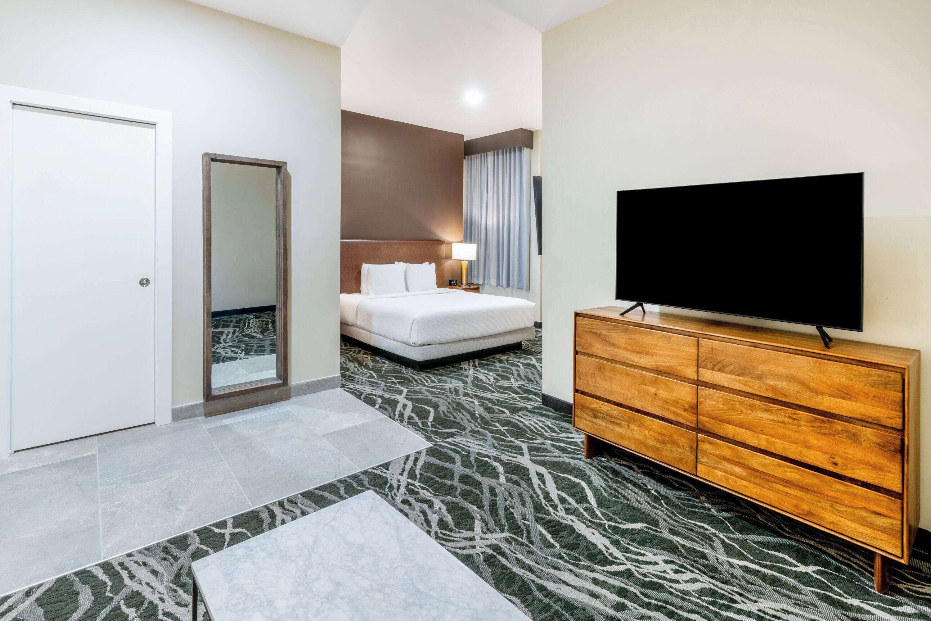 A hotel room with a bed, a television on a wooden dresser, a mirror, and a door.