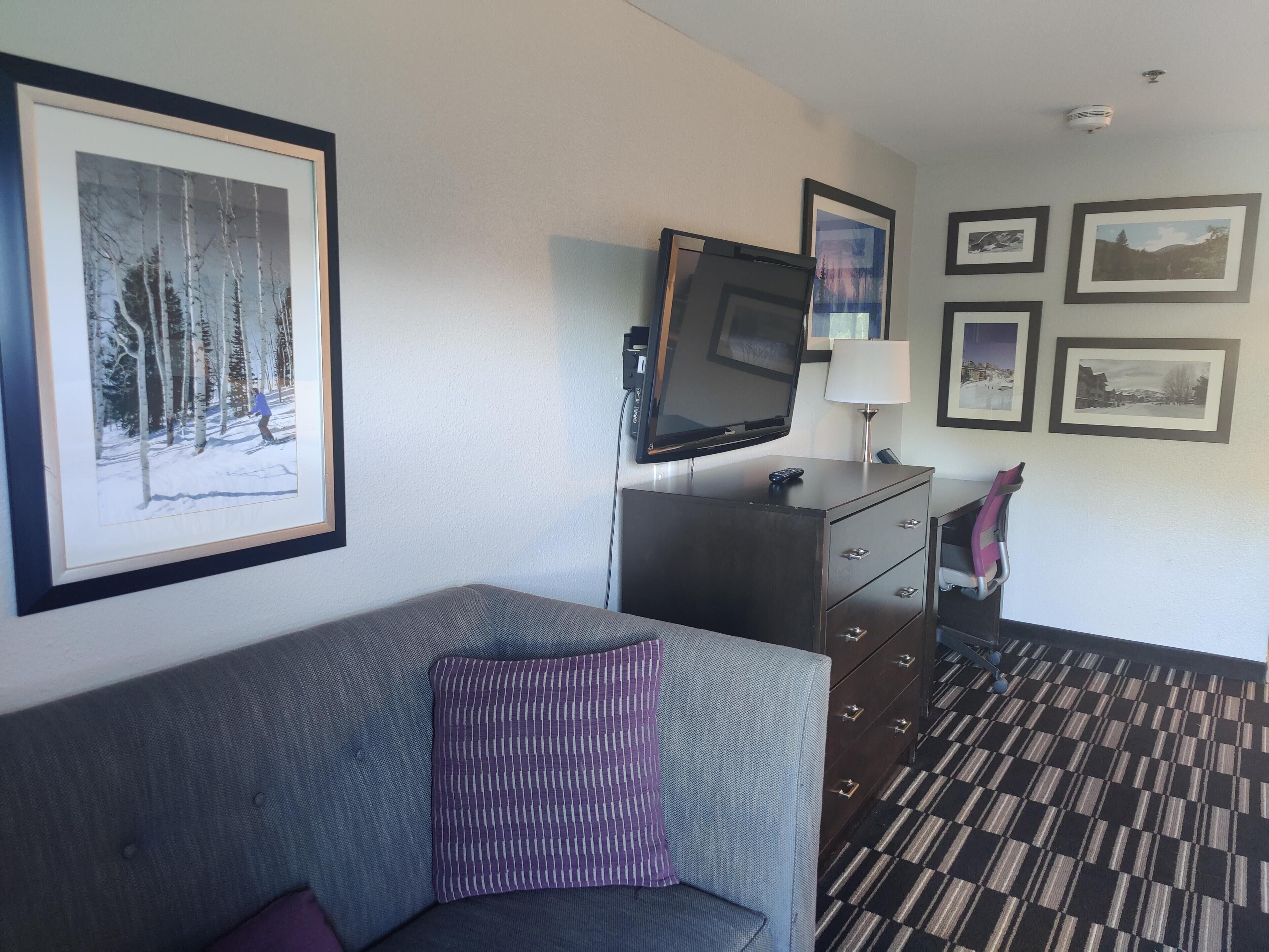 A hotel room with a sofa, a TV, a dresser, and framed pictures on the wall.