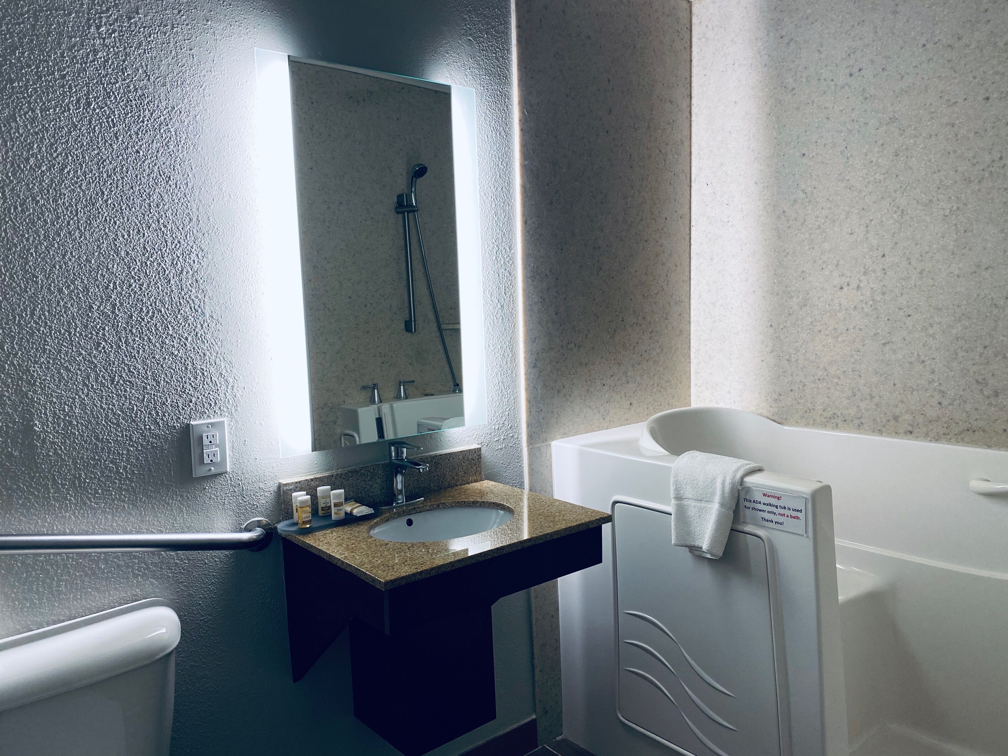 A bathroom with a granite countertop sink, a mirror, a handheld showerhead, and a white bathtub.