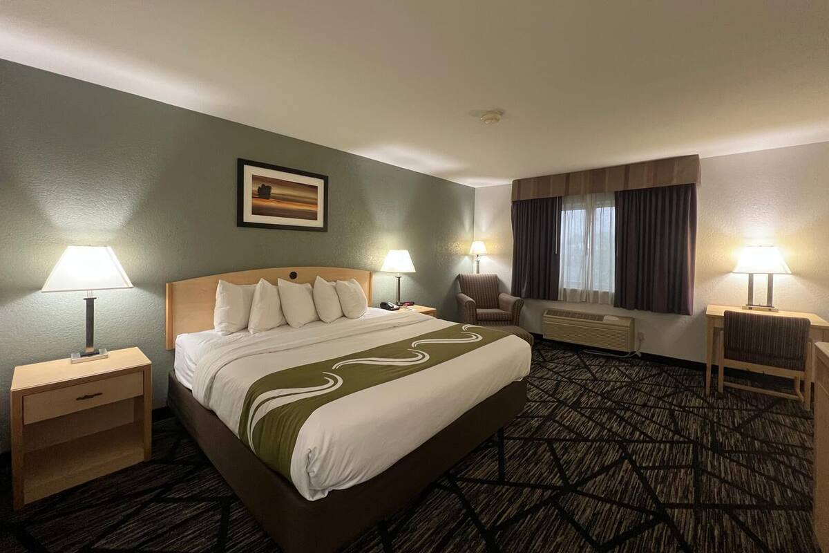 A hotel room with a bed, two bedside tables, a chair, an air conditioning unit, and a window with curtains.