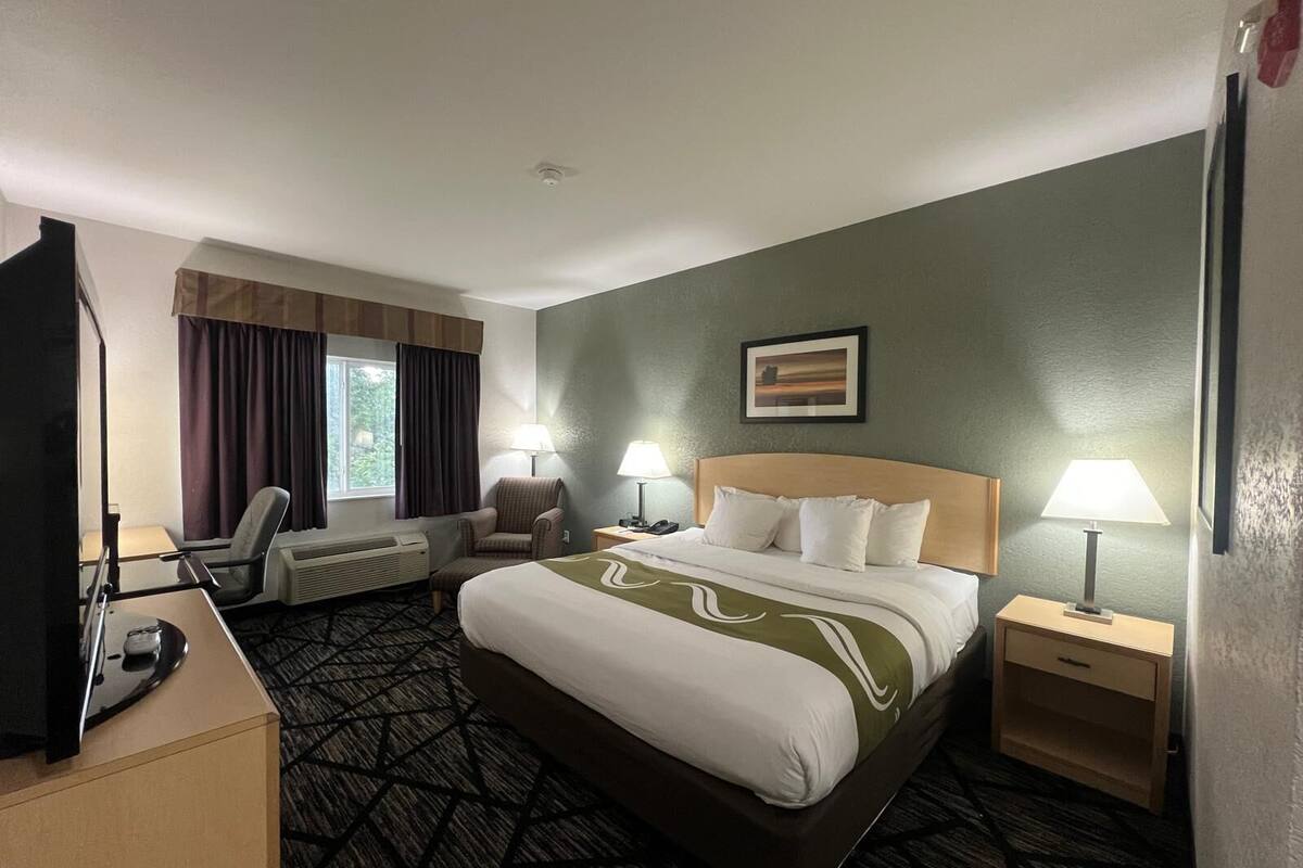 A hotel room with a large bed, two bedside tables with lamps, a chair, a desk with a flat-screen TV, and a window with curtains.