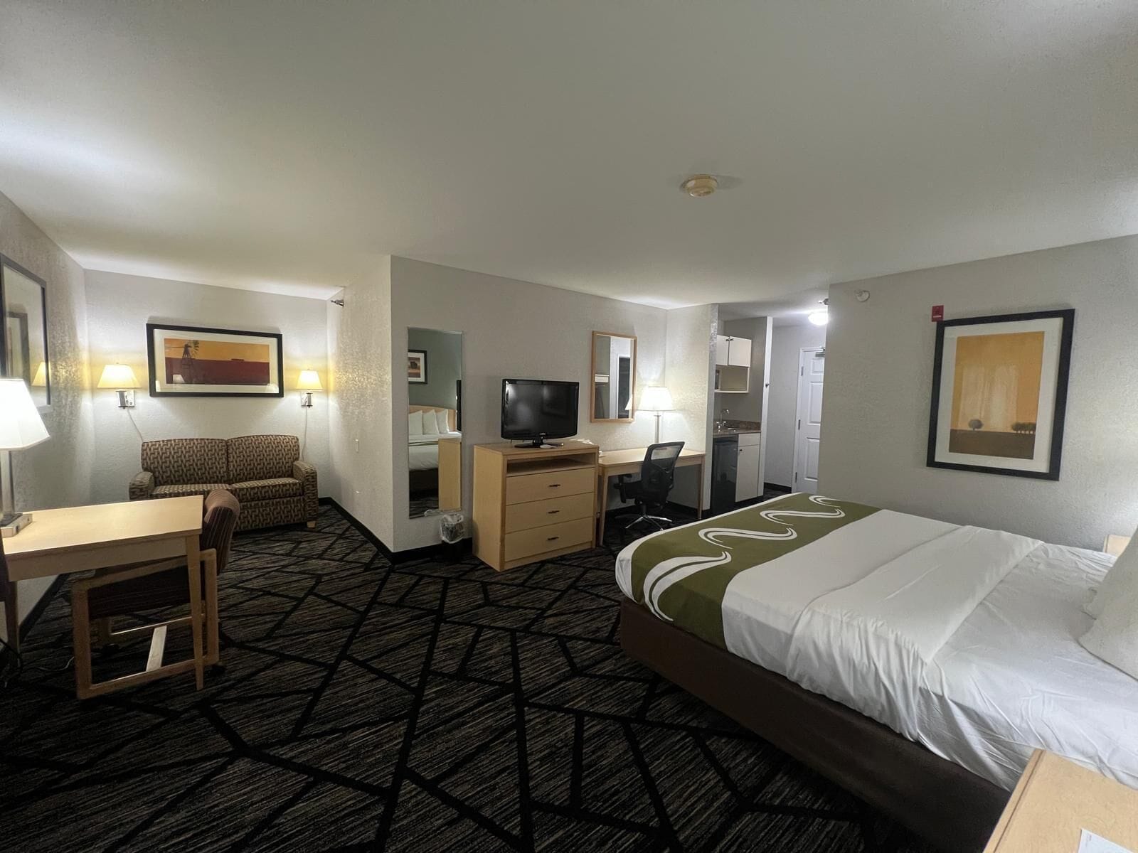 Habitación de hotel con cama, escritorio, silla, televisión y una pequeña cocina.
