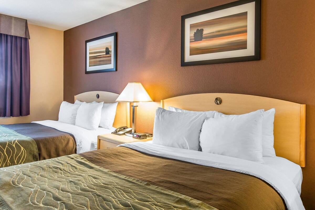 A hotel room with two beds, a bedside table with a lamp, a clock, and framed pictures on the wall.