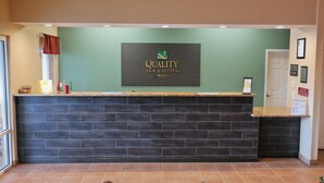 Un comptoir d’accueil avec un revêtement de sol foncé, une enseigne au-dessus indiquant « Quality Inn & Suites, hôtels du groupe Choice Hotels », et une porte sur la droite.