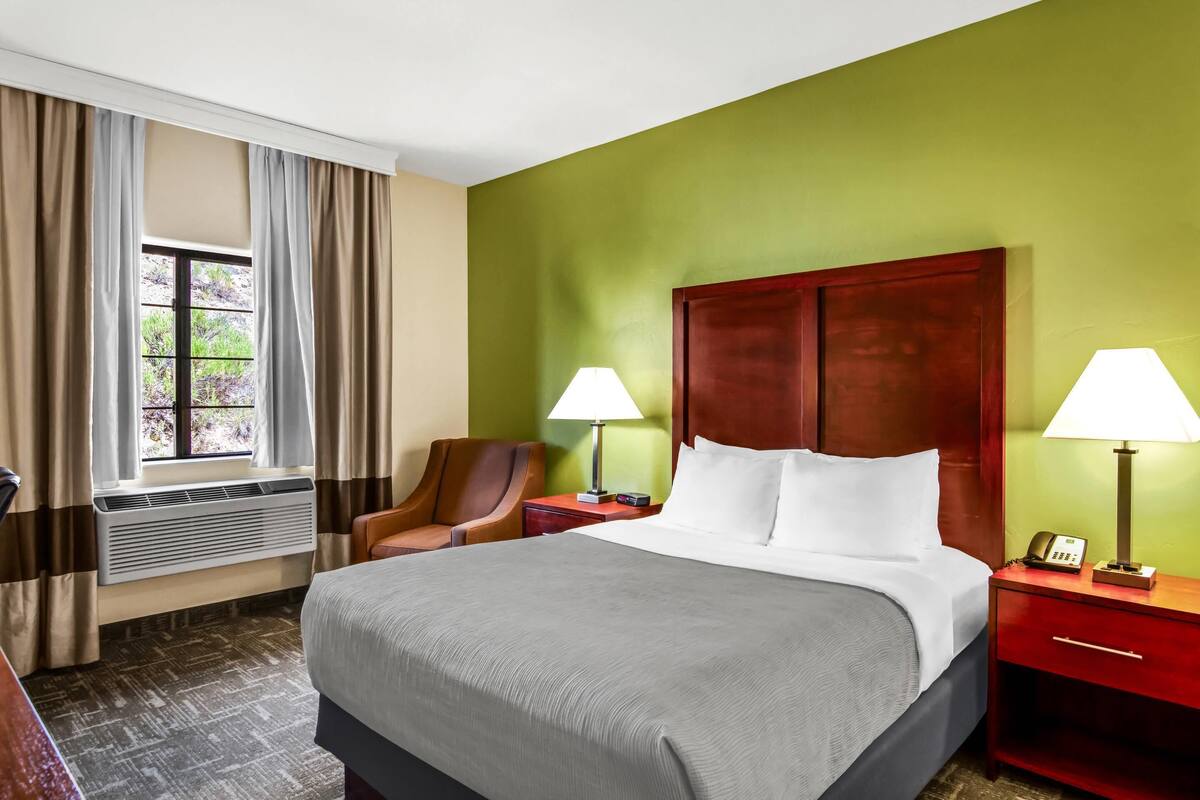 A hotel room with a large bed, two bedside lamps, a wooden headboard, a chair, a nightstand, and a window with curtains.
