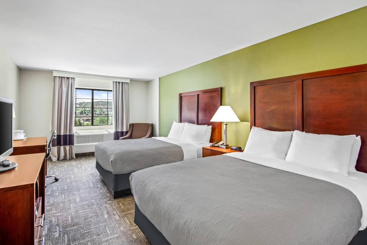 A hotel room with two beds, a desk with a computer, a lamp, and a view of the outdoors.