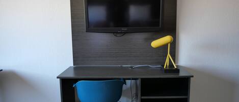 A modern room with a dark wood desk, a blue chair, a mounted flat-screen TV, and a yellow desk lamp.