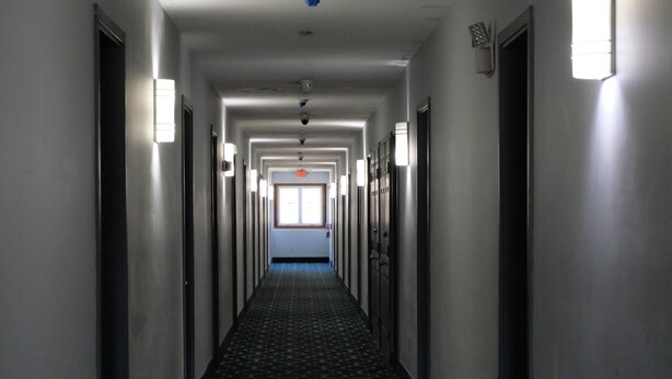 A long, narrow hallway with patterned carpet, doors on both sides, and overhead lighting.