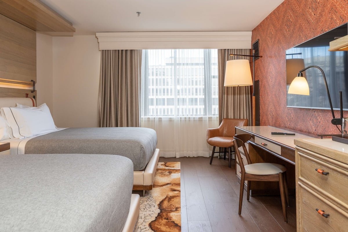 A hotel room with two beds, a desk, a chair, a mirror, and a window with curtains.