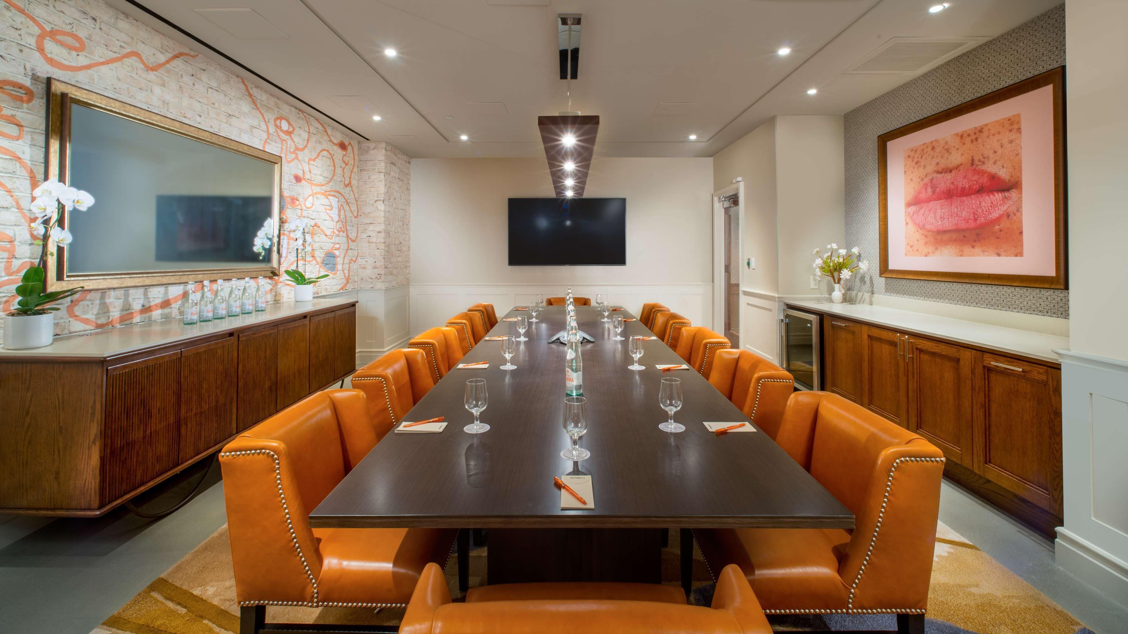 A conference room with a long table, orange leather chairs, a large TV, and decorative elements like orchids and a framed picture.
