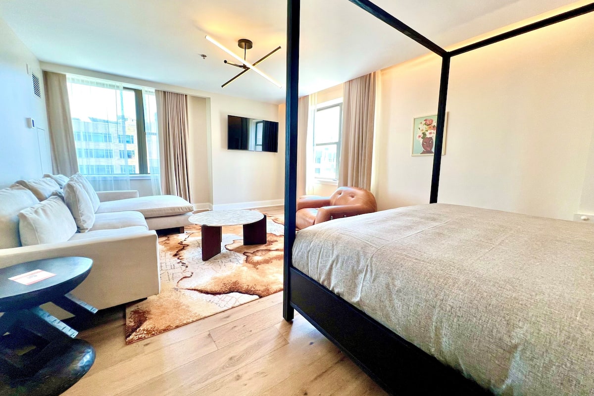 A modern hotel room with a four-poster bed, a sofa, a coffee table, and a TV.