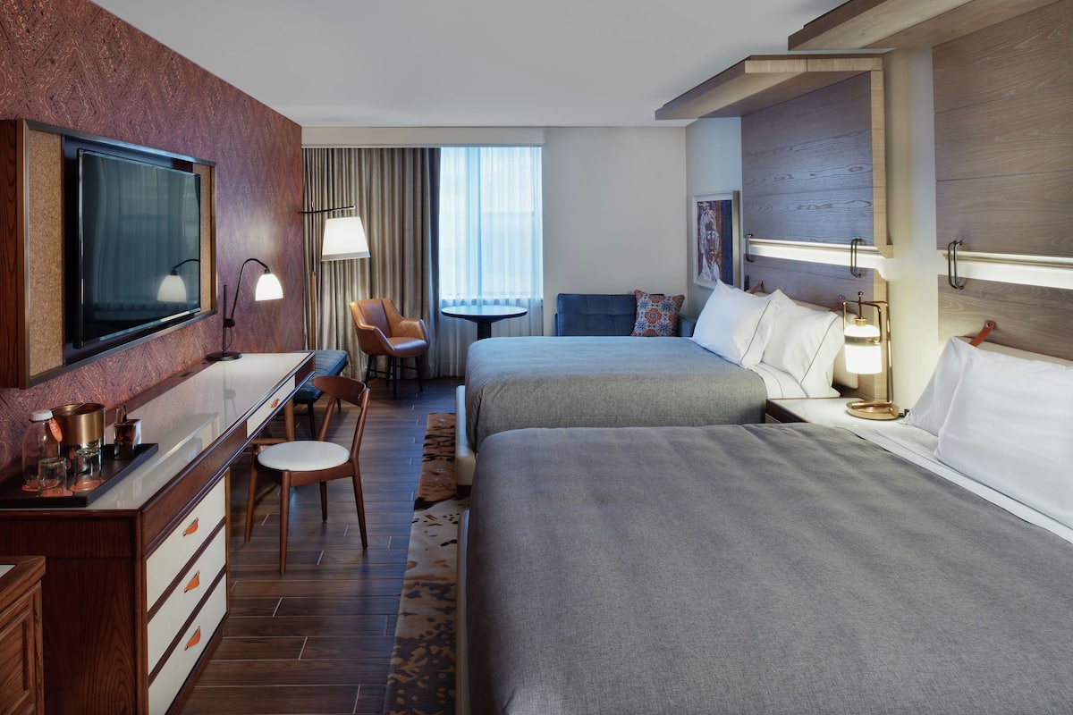 A hotel room with a TV, a desk with a lamp, two beds, a chair, and a small table.