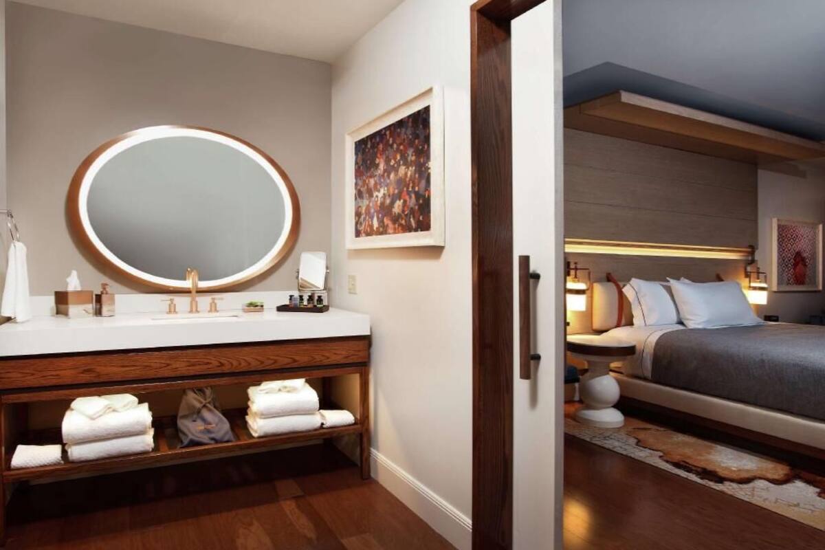 A modern bathroom with a round mirror, wooden vanity, and a bed with a headboard in the adjacent room.