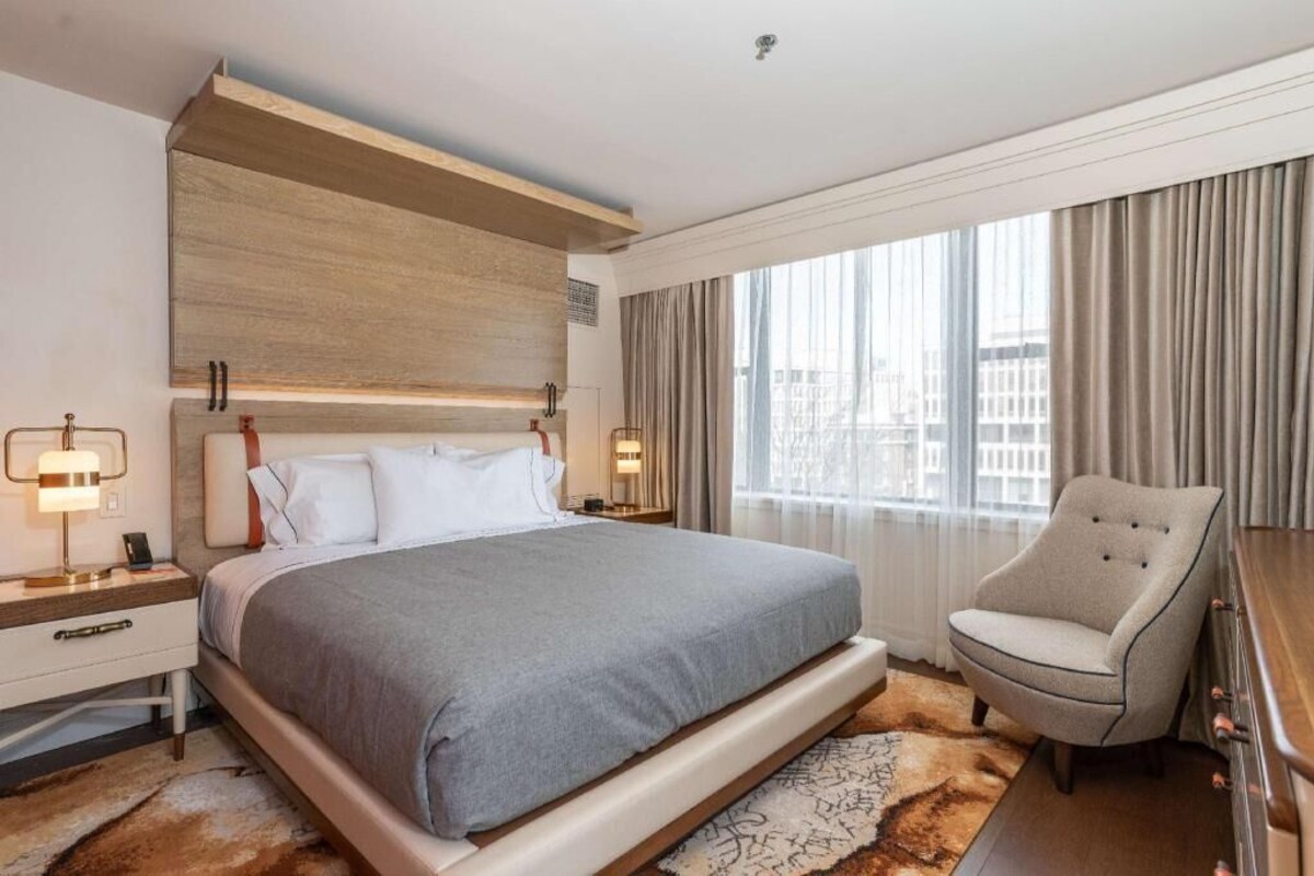A hotel room with a large bed, bedside tables, a chair, a window with curtains, and a city view.