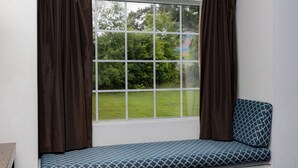 Ein Fenster mit braunen Vorhängen, Blick ins Grüne und eine blau gemusterte Bank.