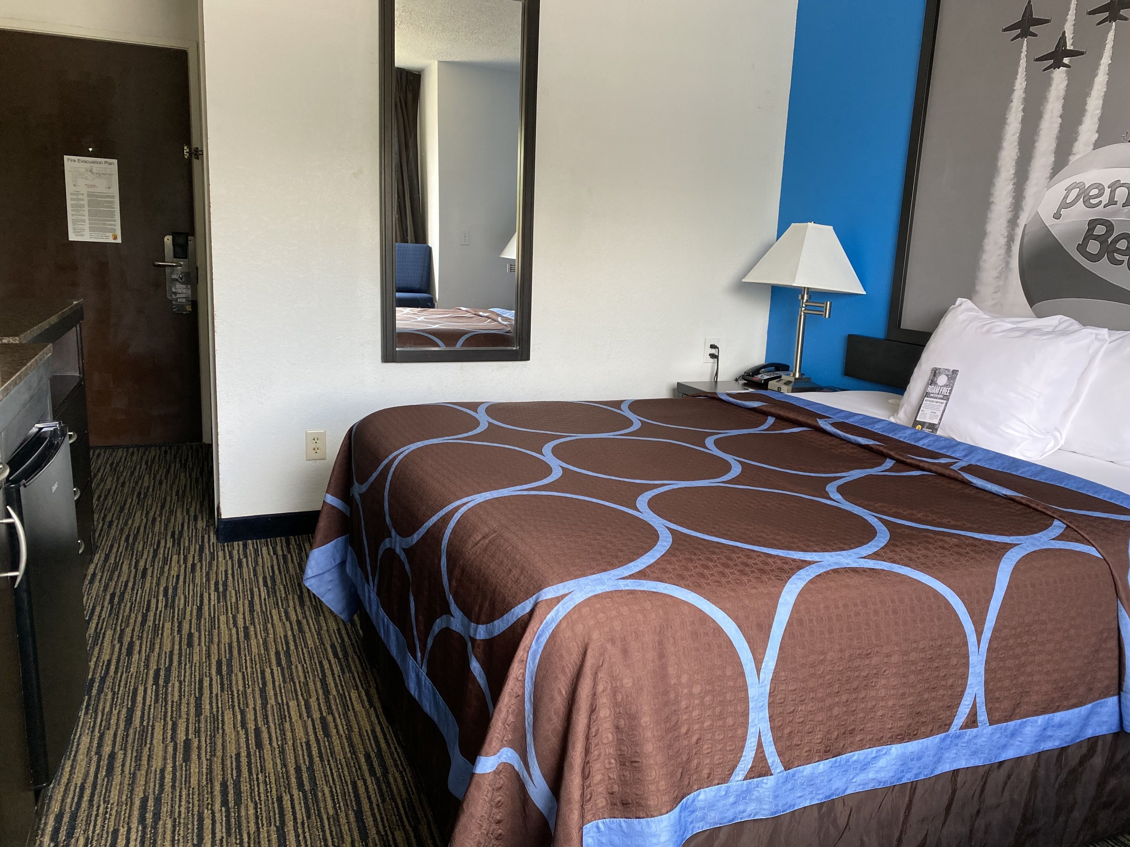 A hotel room with a bed, a desk, a mirror, a lamp, and a wall with a poster.
