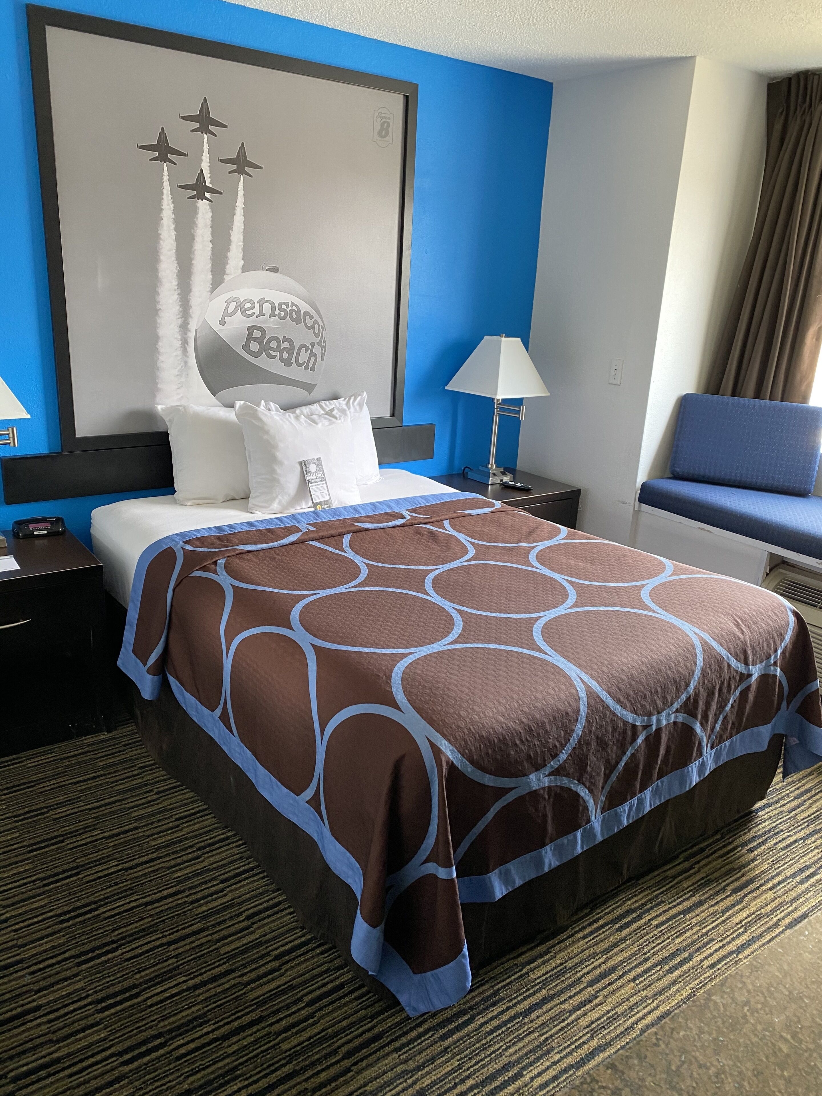 A hotel room with a bed, a nightstand, a lamp, a chair, and a wall with a poster.
