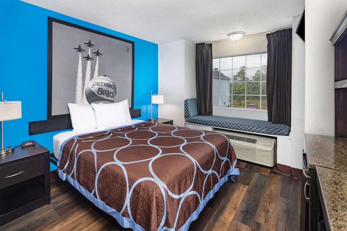 Habitación de hotel con cama, asiento junto a la ventana, una pared decorada con arte temático de playa y mesita de noche con lámpara.