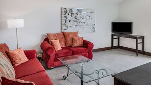 Ein Wohnzimmer mit einem roten Sofa, einem Glastisch, einem Fernseher auf einem Schrank und einem Bild an der Wand.