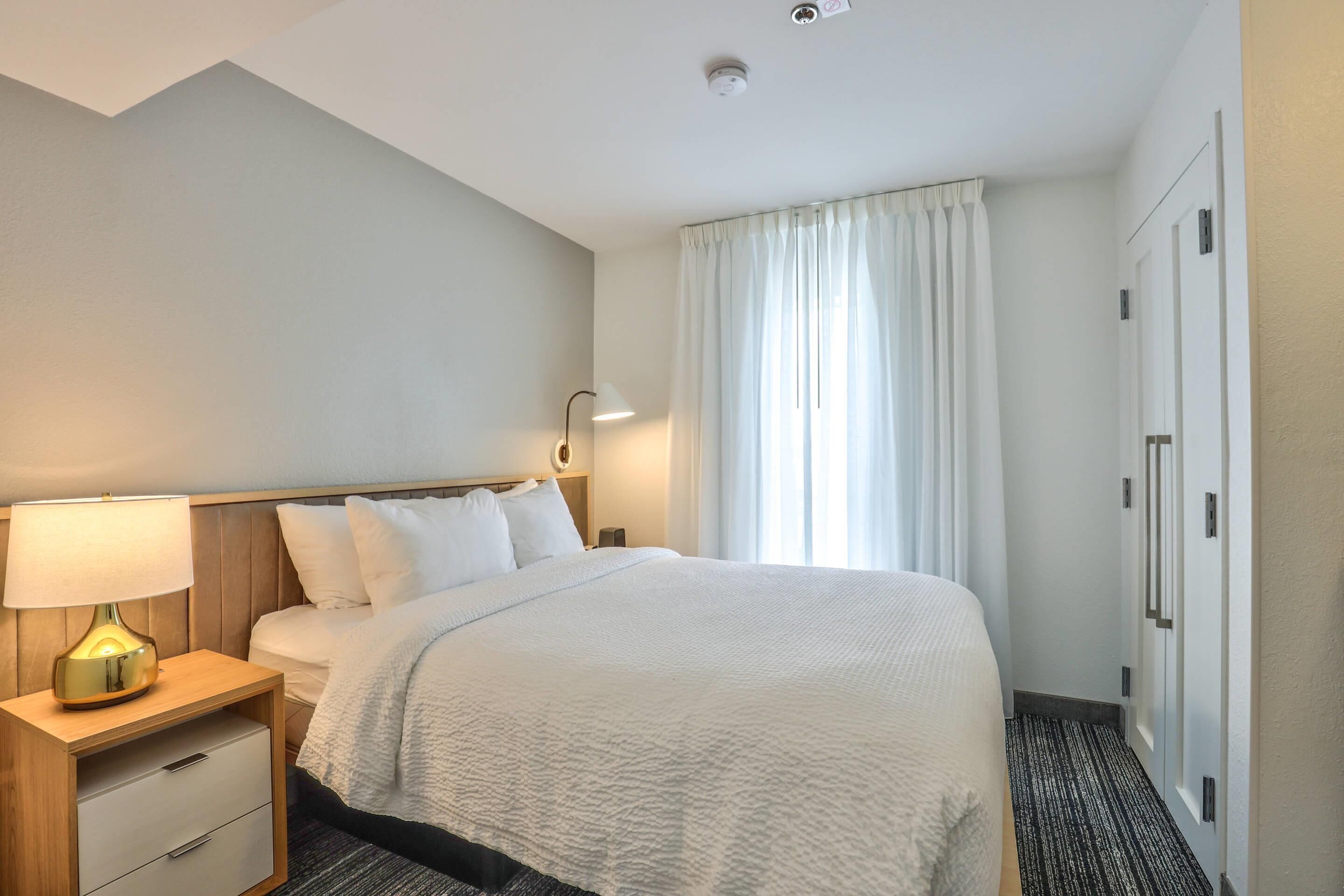 Quarto de hotel com cama, mesa de cabeceira com abajur, guarda-roupa e janela com cortinas.