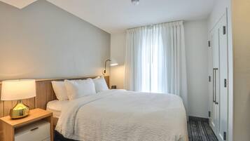 Quarto de hotel com cama, mesa de cabeceira com abajur, guarda-roupa e janela com cortinas.