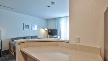 A modern hotel room with a curved beige counter, a gray sofa, a flat-screen TV, and a window with sheer curtains.