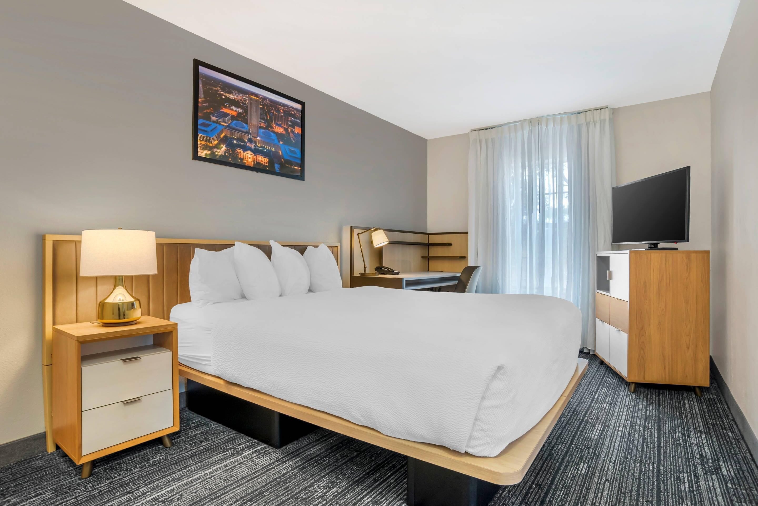 A hotel room with a large bed, a desk, a television, and a cityscape view.